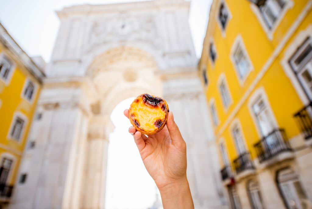 a segurar um pastel de torta de ovo portugues chamado pastel de nata ao ar livre no fundo do arco triunfal em lisboa scaled 1
